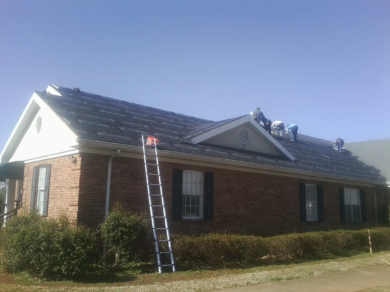 Crew tearing shingles off a brick ranch roof with a ladder set — Emergency Roof Repair work by our crew in Dunnsville