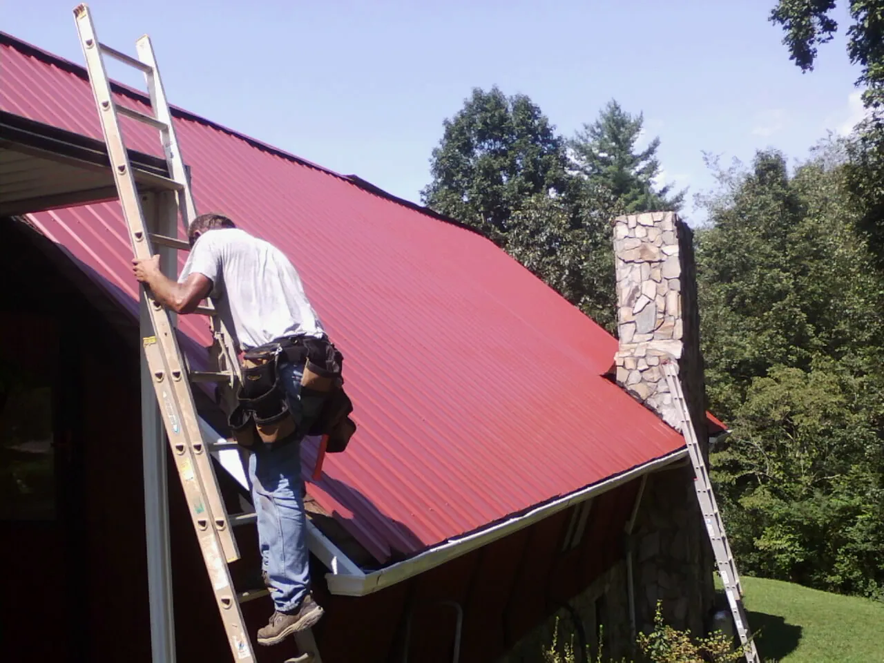 Roofer on a ladder beside a red metal roof and stone chimney — Soffit & Fascia Repair by Roofing Brothers in Dunnsville